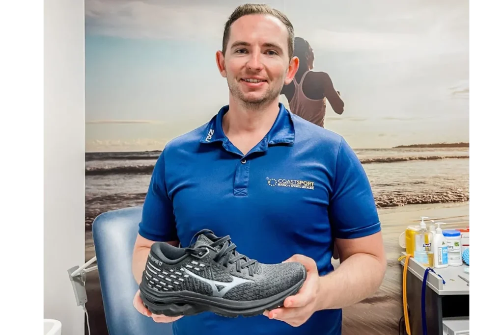 Man holding a black running shoe in front of Coast Sport background.
