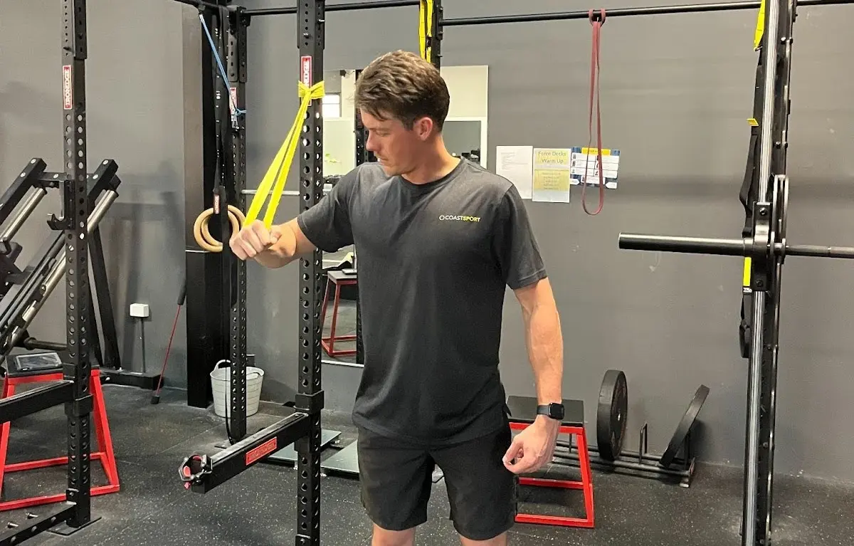 Man performing a shoulder mobility exercise in a gym, using yellow resistance bands attached to a squat rack, focusing on controlled arm movement and posture.